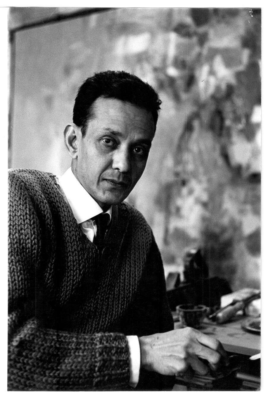 Black and white portrait of S. H. Raza: there's an abstract painting int he background, he sits three-quarters facing the viewer, looking at the camera, his hand upon something